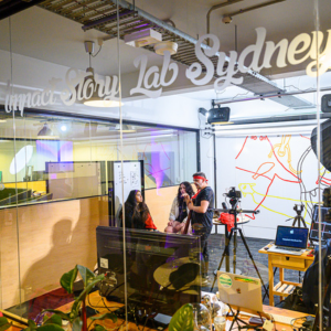 People filming an interview inside a modern studio space, viewed through glass with “Impact Story Lab Sydney” written on it.